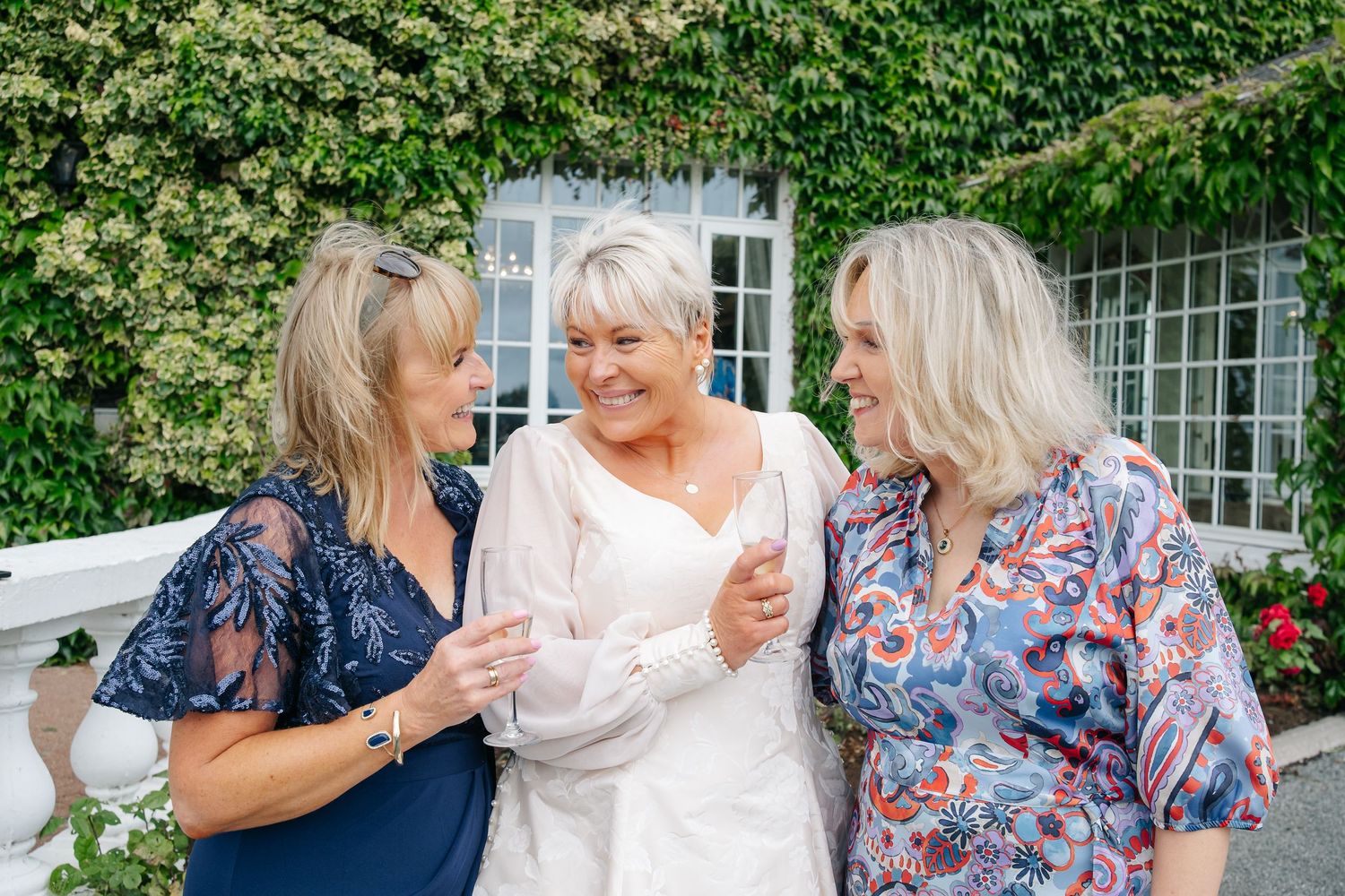 Three women laugh and embrace outdoors in front of an ivy-covered wall at a wedding celebration.