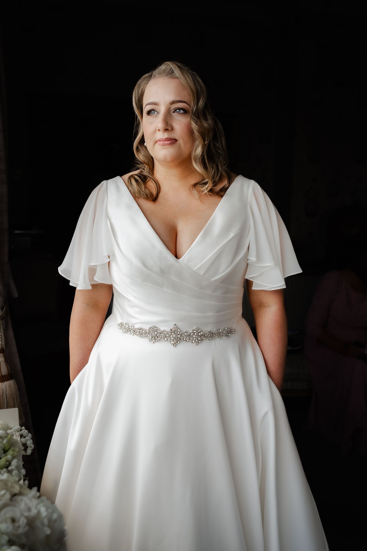 Plus-size bride in white V-neck wedding gown with flutter sleeves and crystal belt, posing against dark background.