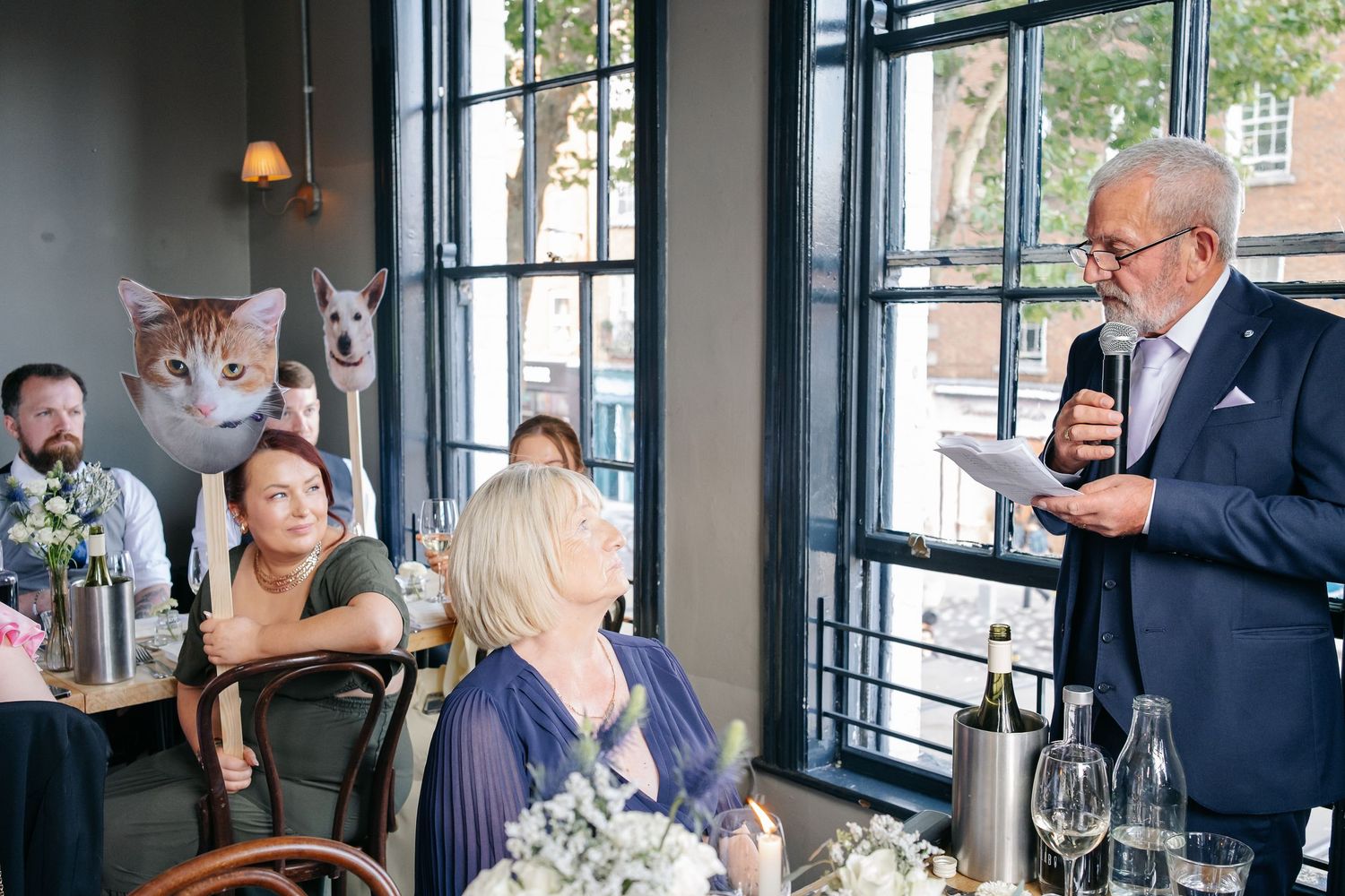 Wedding guests enjoy a stylish reception in a bright restaurant with large windows and elegant table settings.