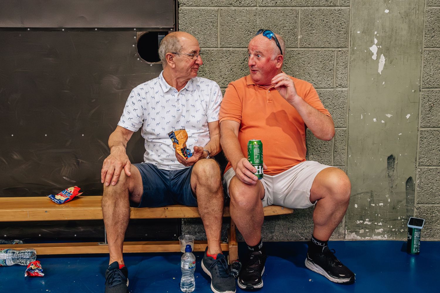 Two people sitting on bench sharing drinks and conversation in gym setting.