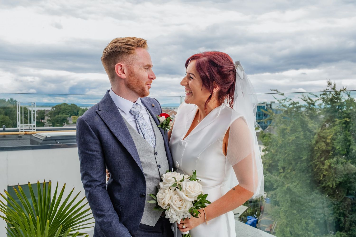 Couple shares intimate moment on balcony with cloudy sky and greenery in background.