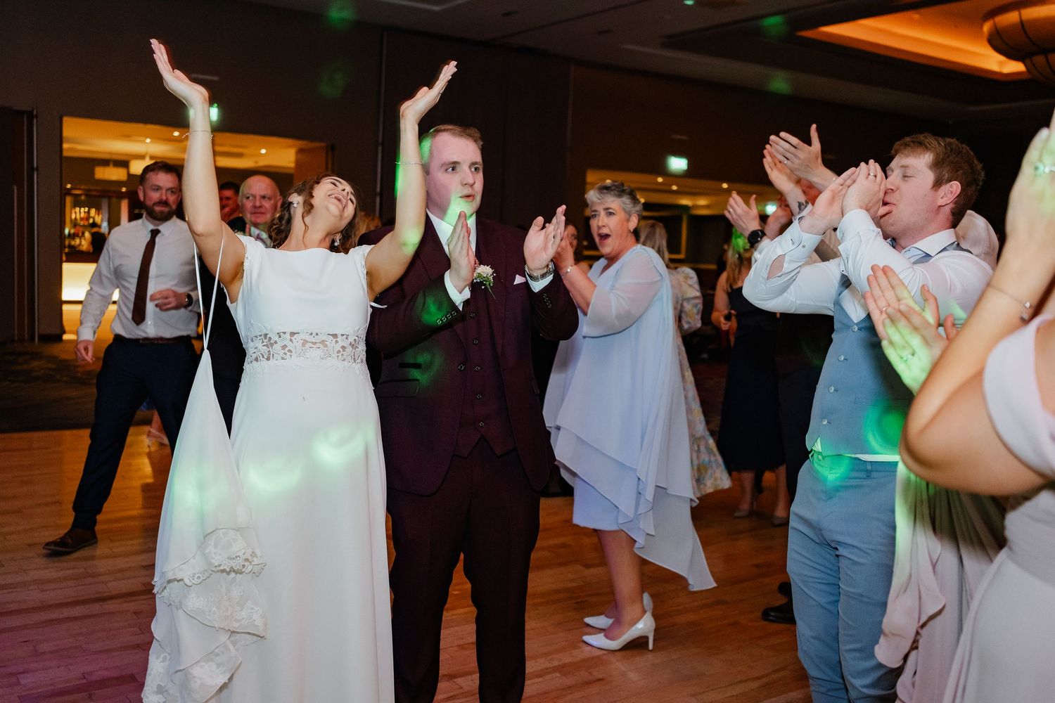 Wedding guests dance and celebrate on a wooden dance floor under colorful lighting at an indoor reception venue.