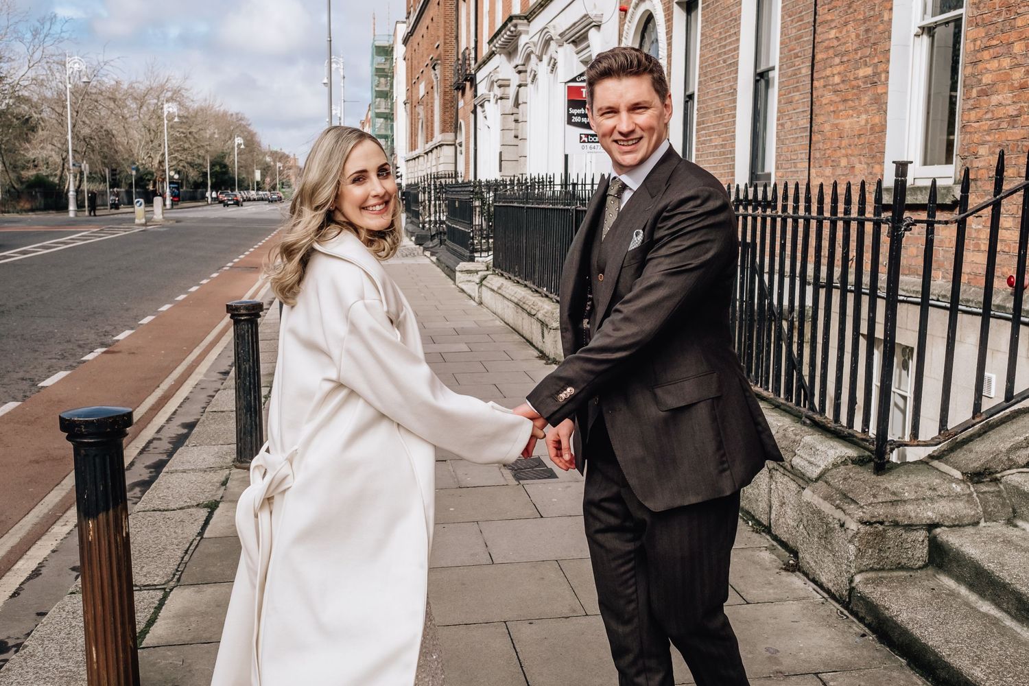 Newlyweds hold hands and smile while posing on a city sidewalk in winter attire.