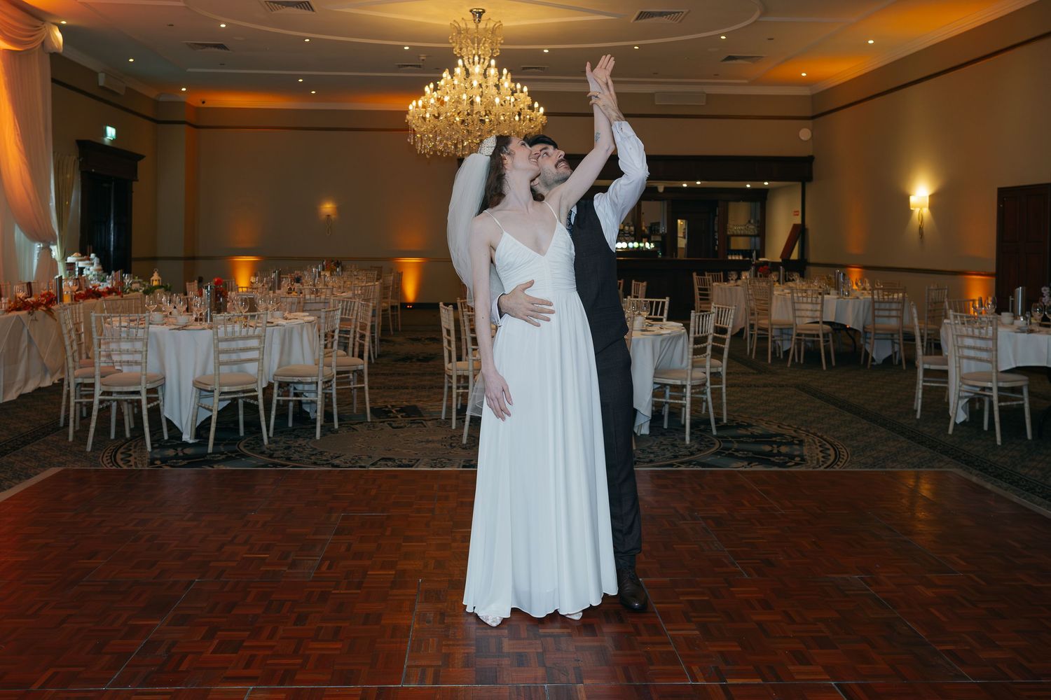 Elegant ballroom venue with crystal chandeliers and dark wood dance floor awaits wedding celebration.
