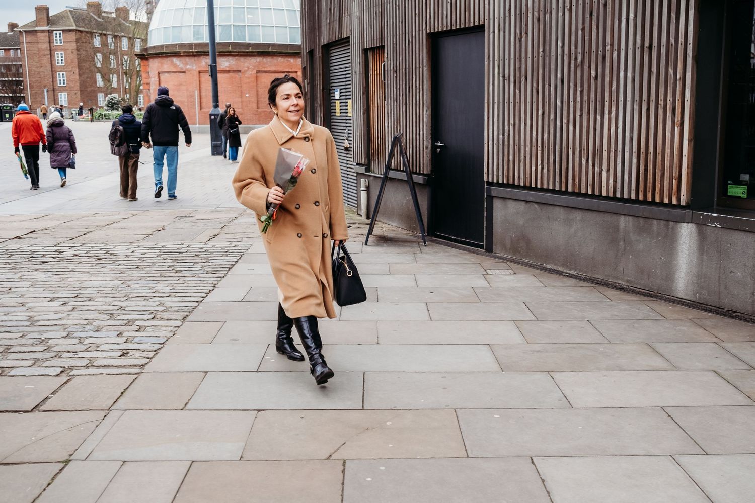 Person in tan coat walking on cobblestone street in urban setting.