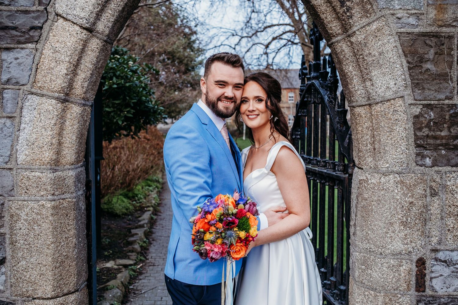 A couple in wedding attire poses under a stone archway holding a vibrant bouquet.