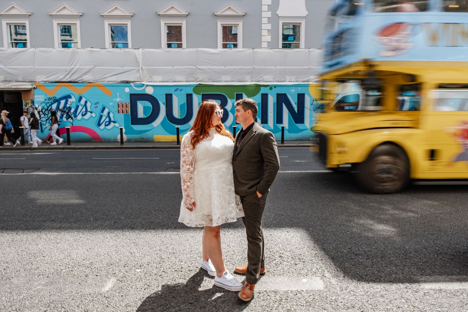 A couple poses in front of a vibrant Dublin street mural with a yellow school bus passing by.