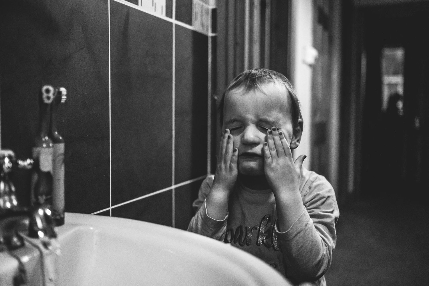 Black and white photo showing child washing face at bathroom sink.