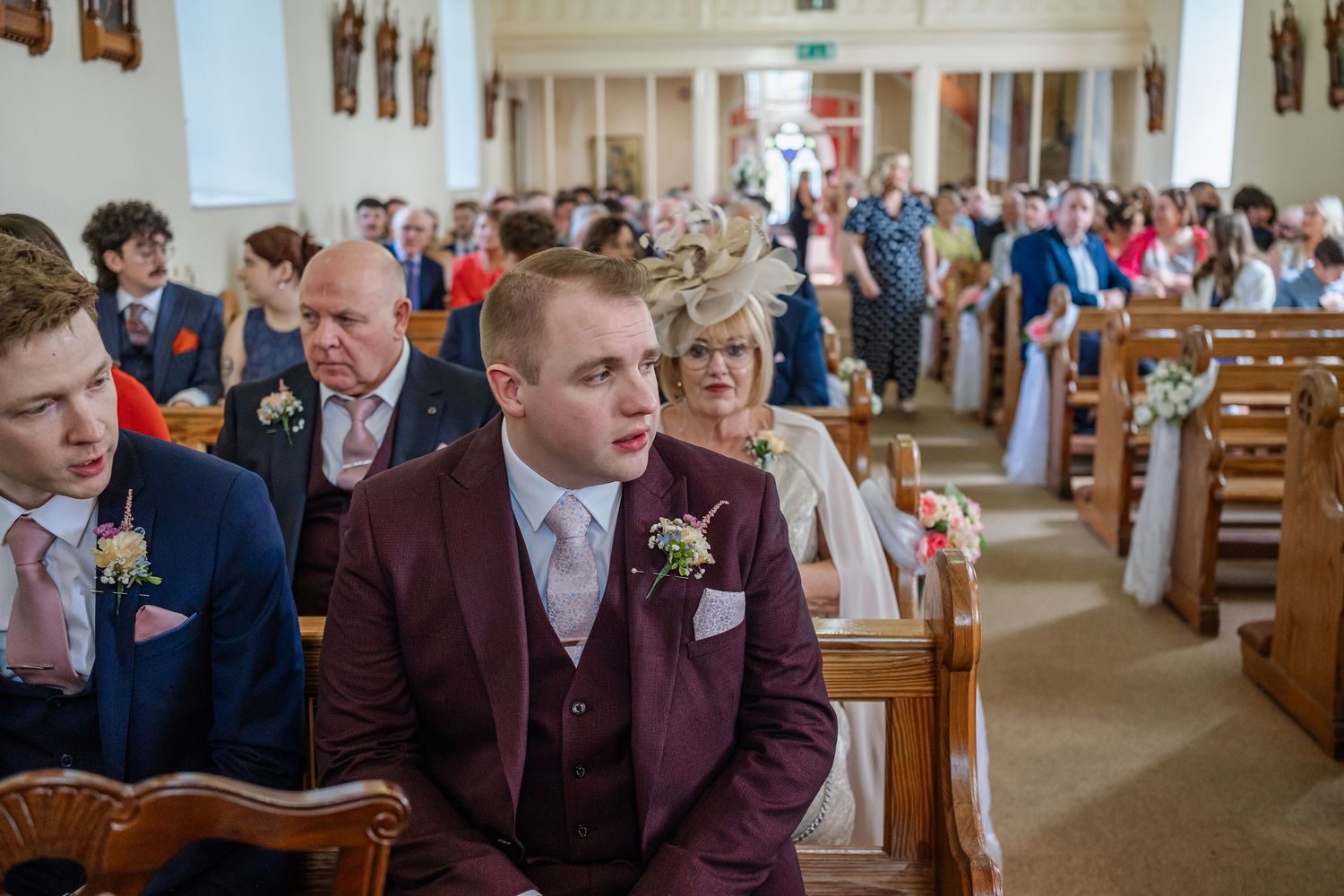 A formal church ceremony with guests seated in wooden pews inside an elegant white-walled sanctuary.