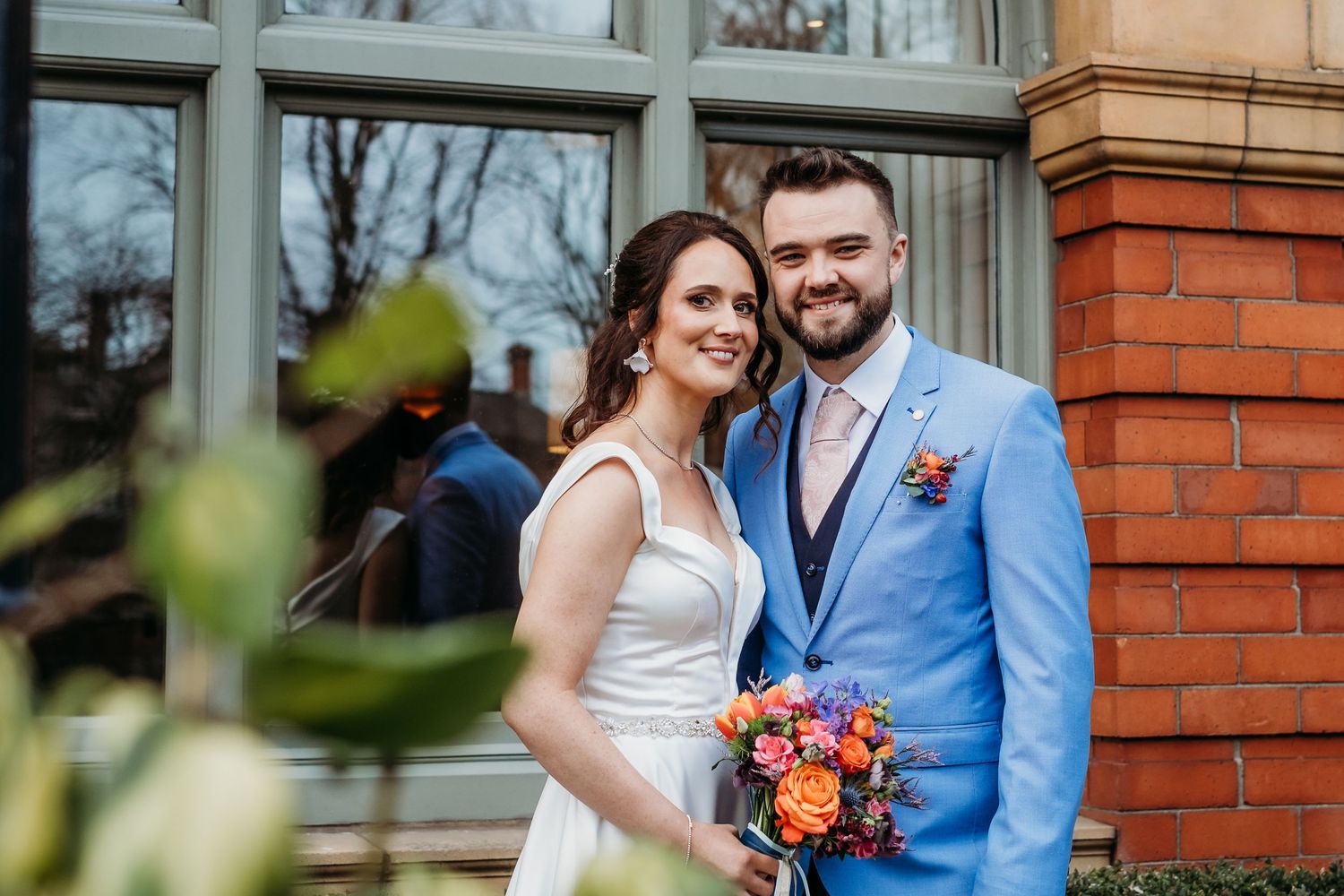 Wedding couple poses in front of red brick building wearing blue suit and white dress.
