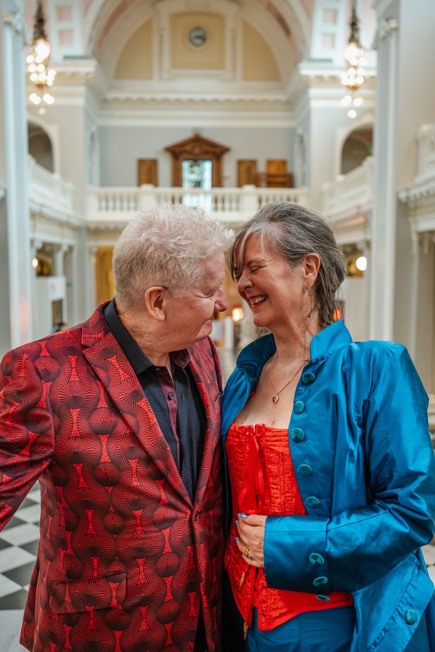 A couple sharing an intimate moment in an elegant hall with ornate architecture and chandeliers.