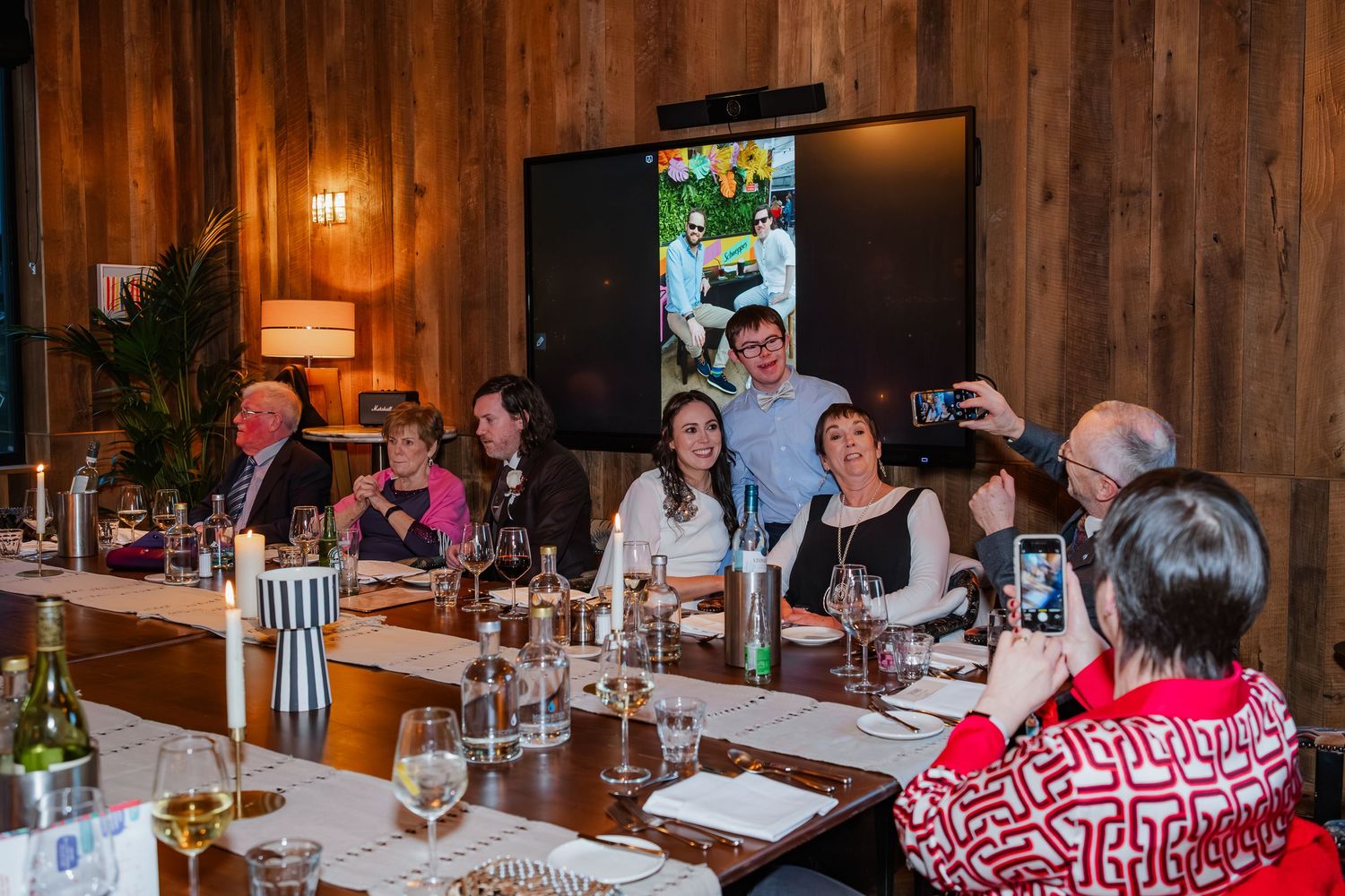 Group of guests smiling at dinner table during wedding reception with wine glasses and TV screen.