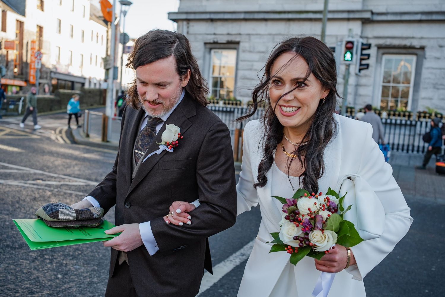 Bride holding bouquet and groom with green folder walking on city street after wedding ceremony.