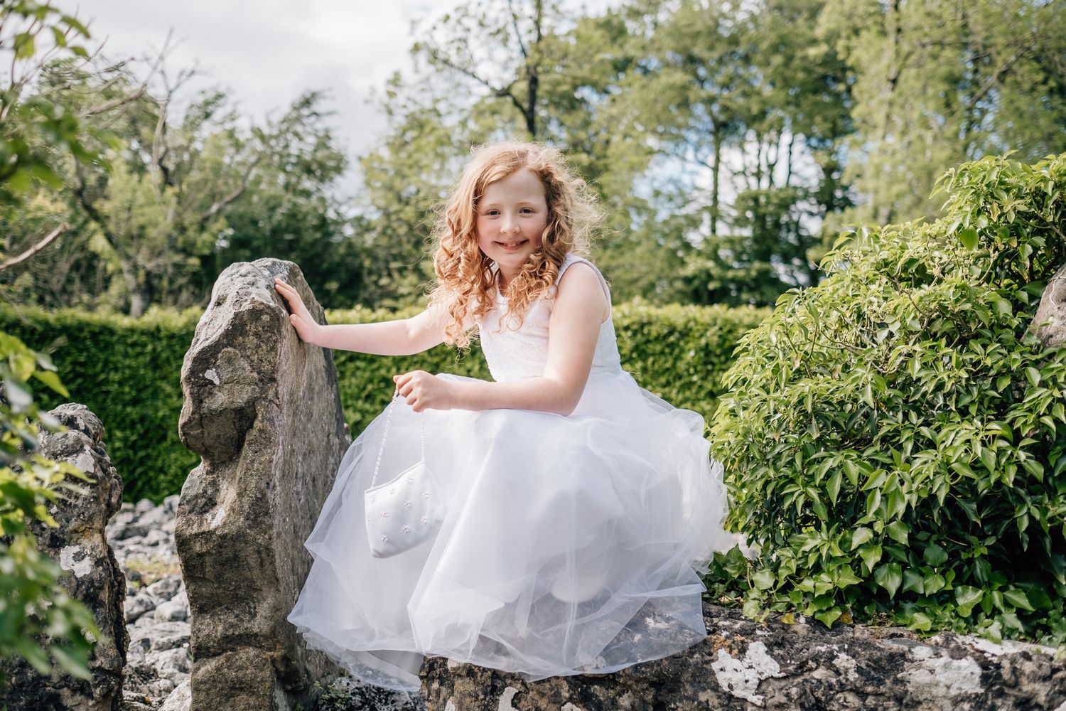 Young girl in white dress posing on stone wall in lush garden setting.