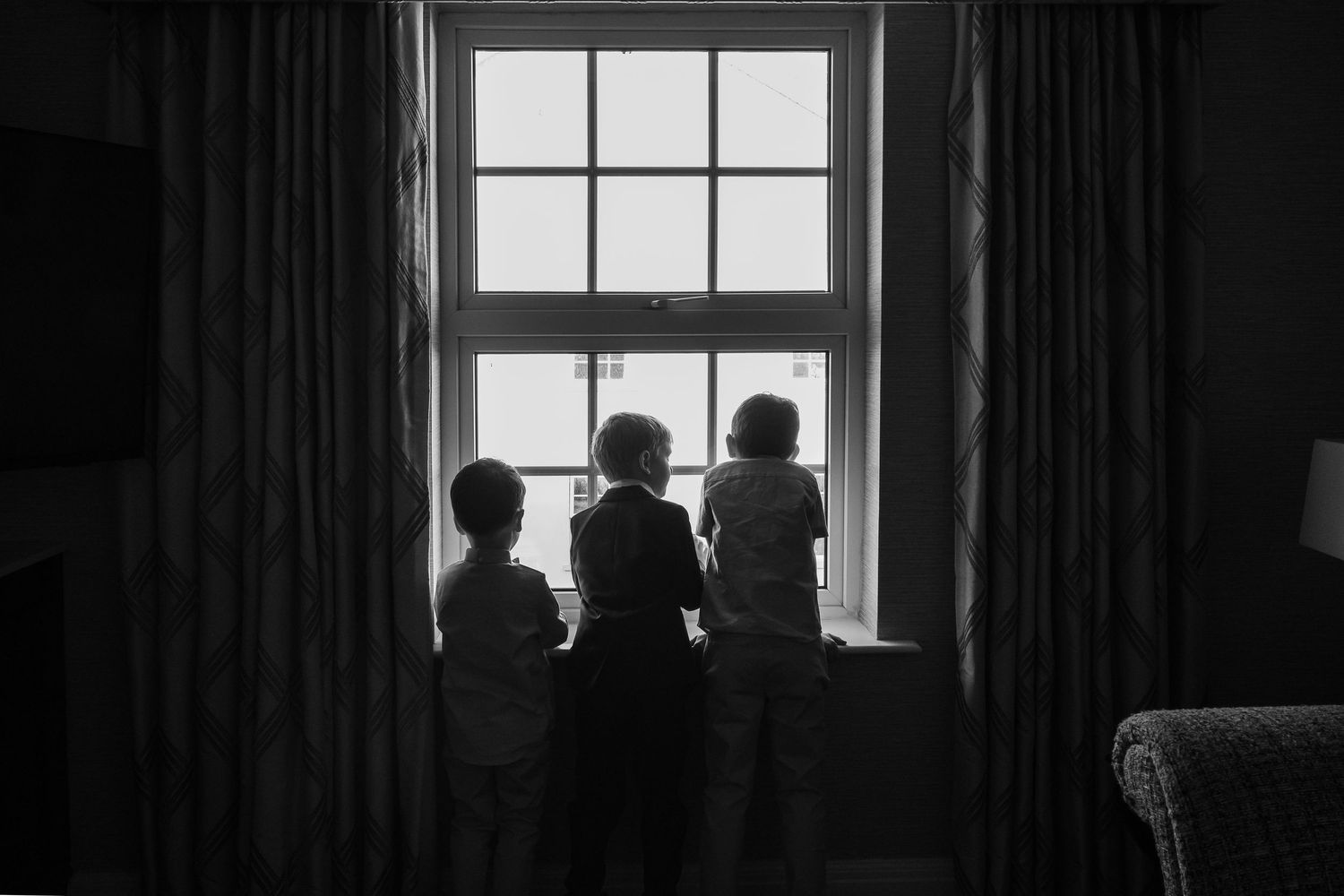 Three children silhouetted against a bright window, looking outside in a black and white photo.