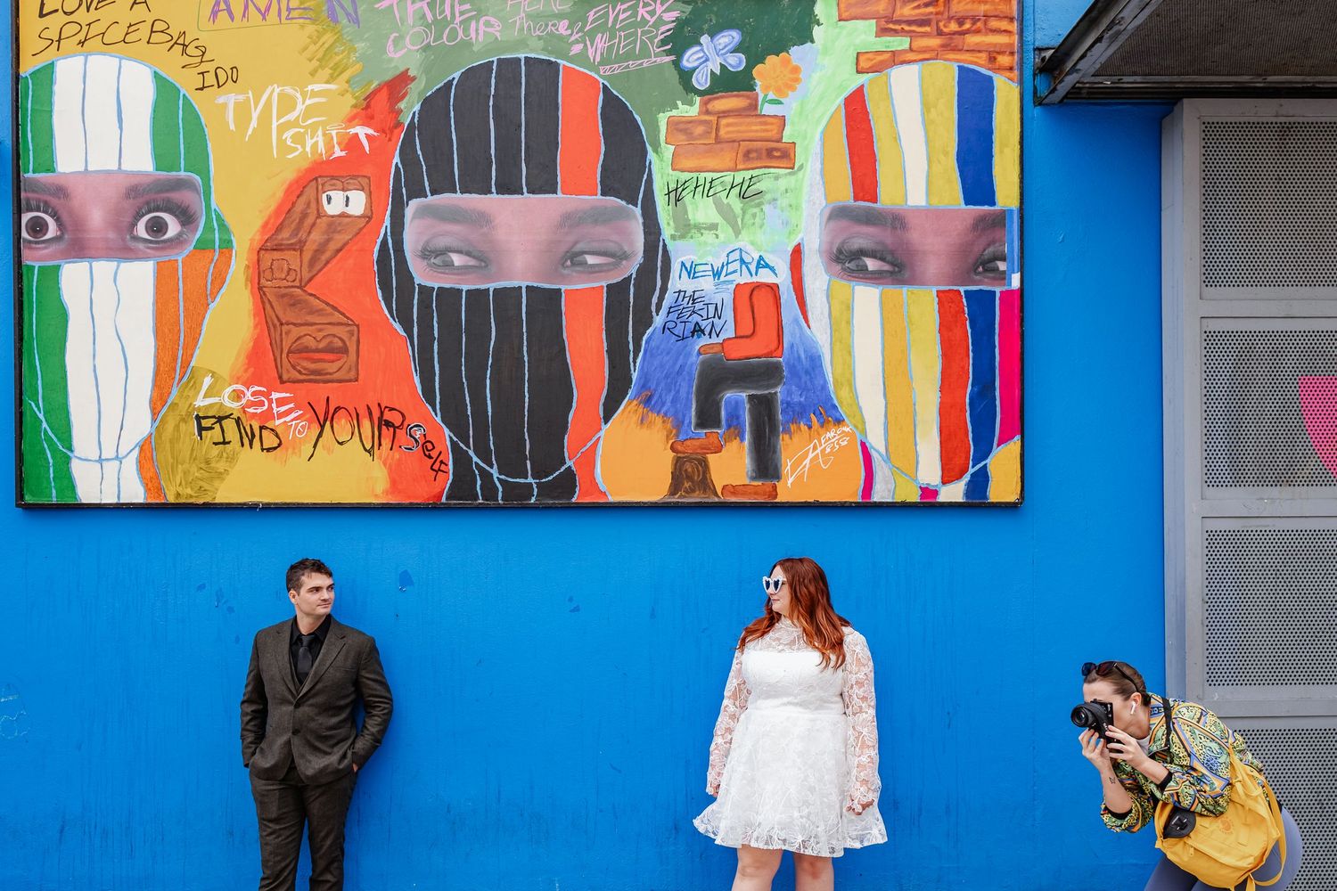 Couple poses against bright blue wall with colorful artistic murals creating playful wedding portrait.