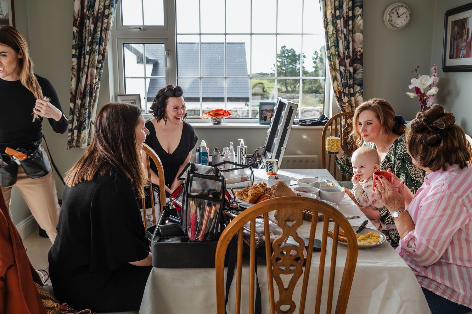 Women gathered around a makeup station in a bright dining room during bridal preparations.