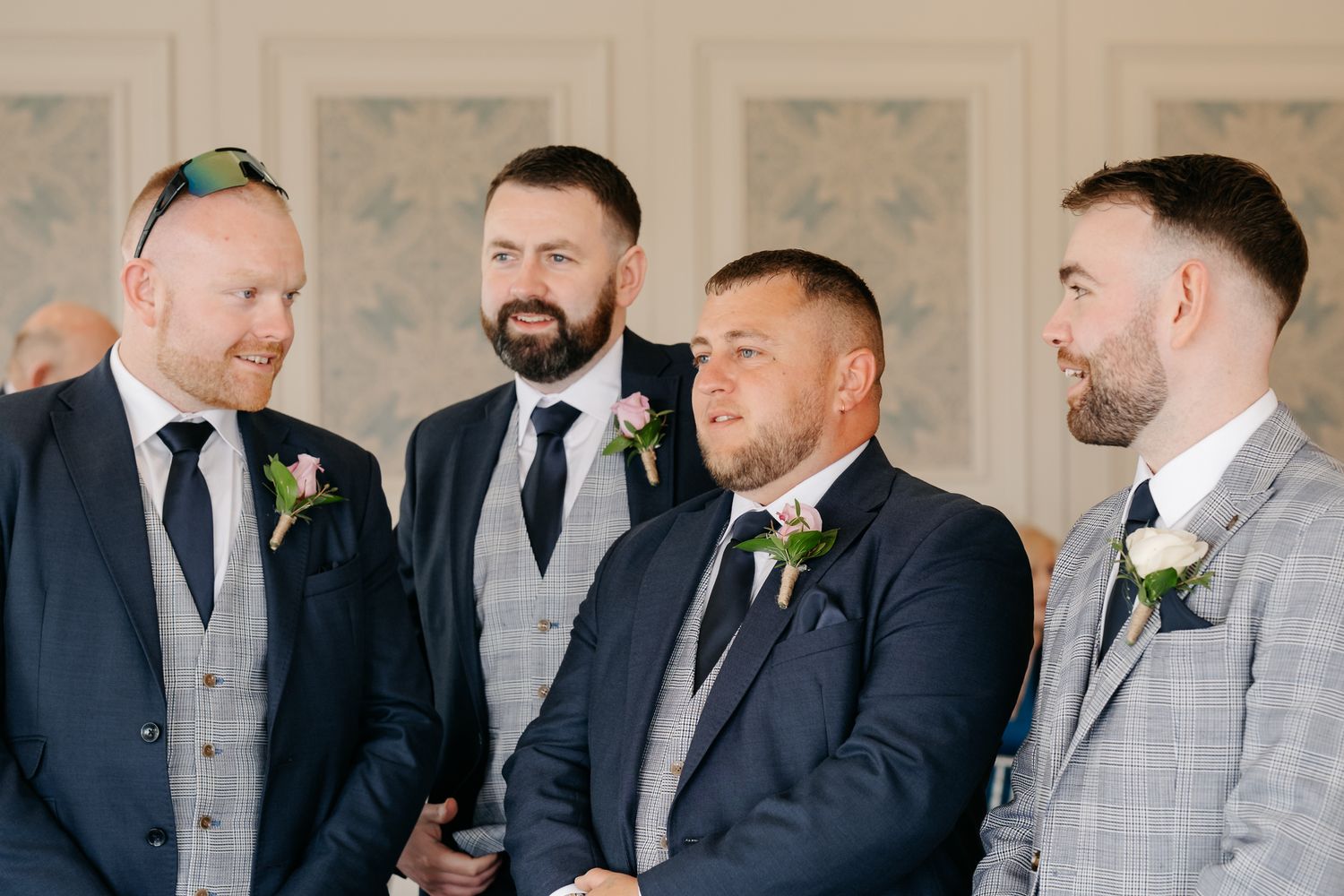 Four groomsmen in navy and grey suits with floral boutonnieres smiling during a wedding ceremony.