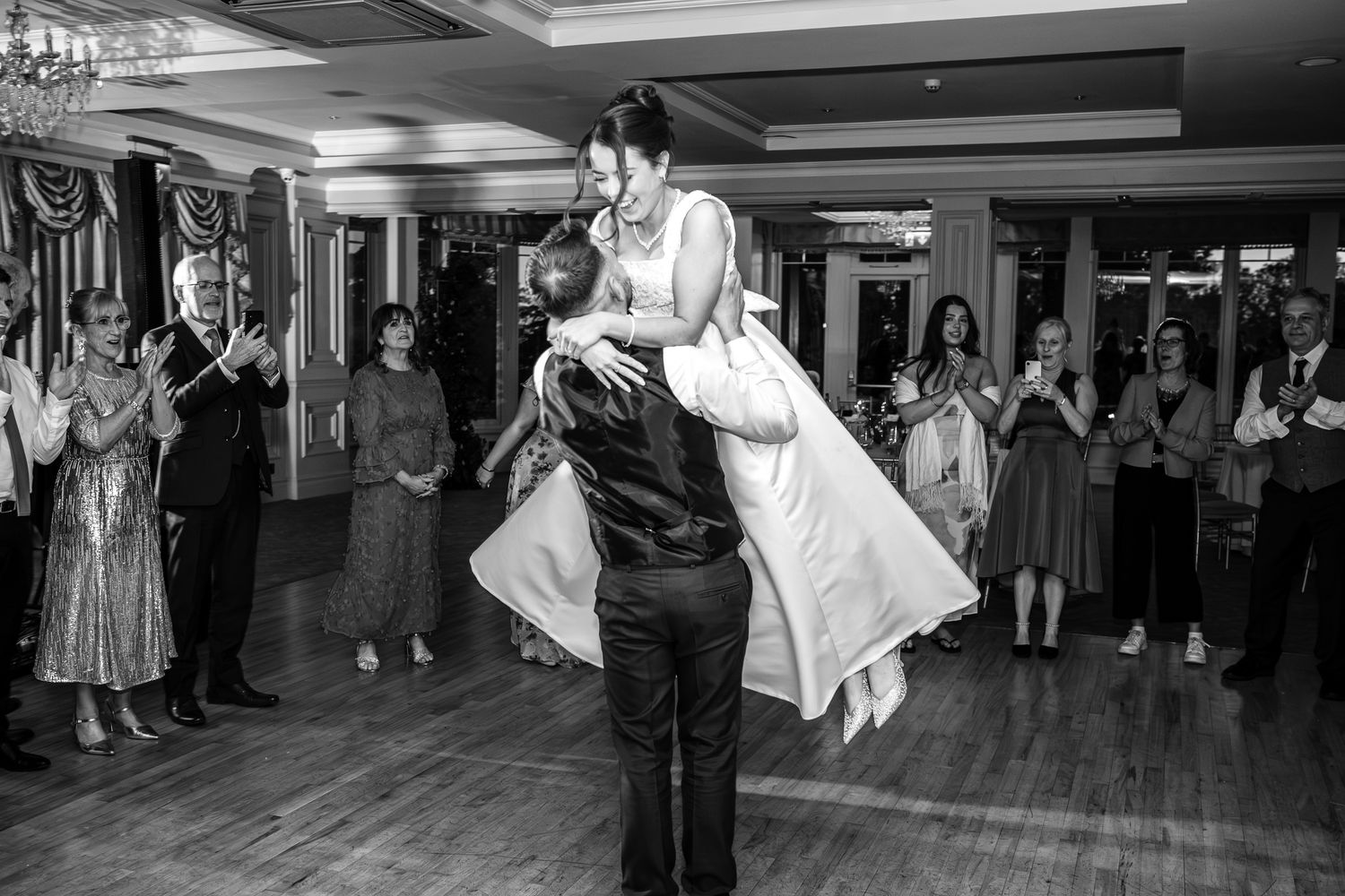 Groom lifts bride during first dance at wedding reception as guests clap and celebrate in black and white photo.