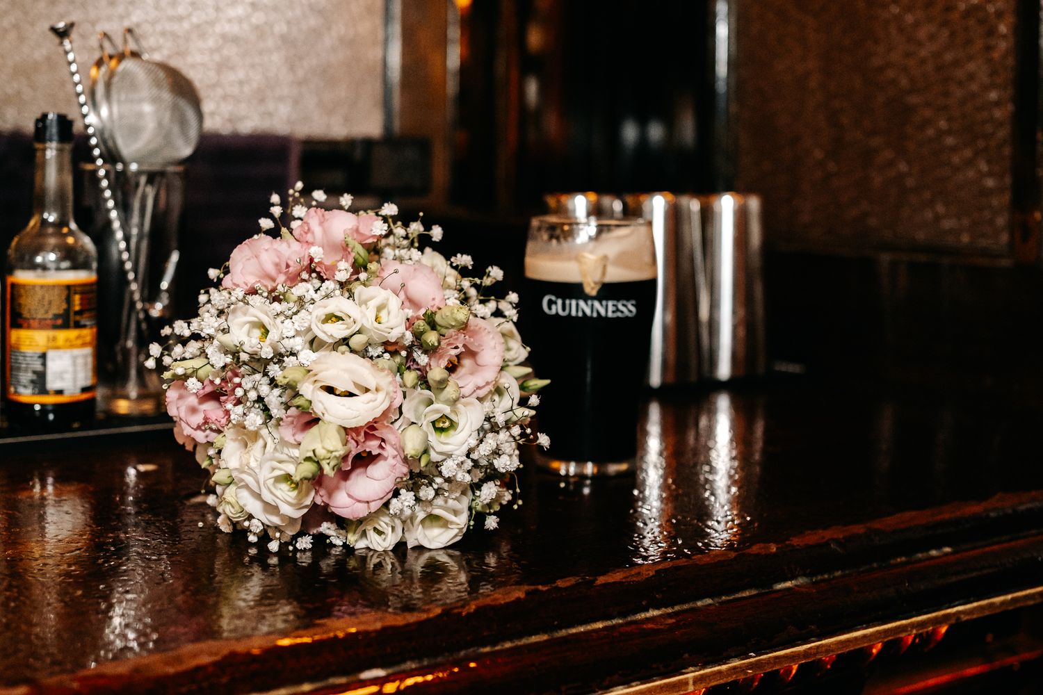 Wedding bouquet of pink roses and baby's breath beside a pint of Guinness on a dark wooden bar.