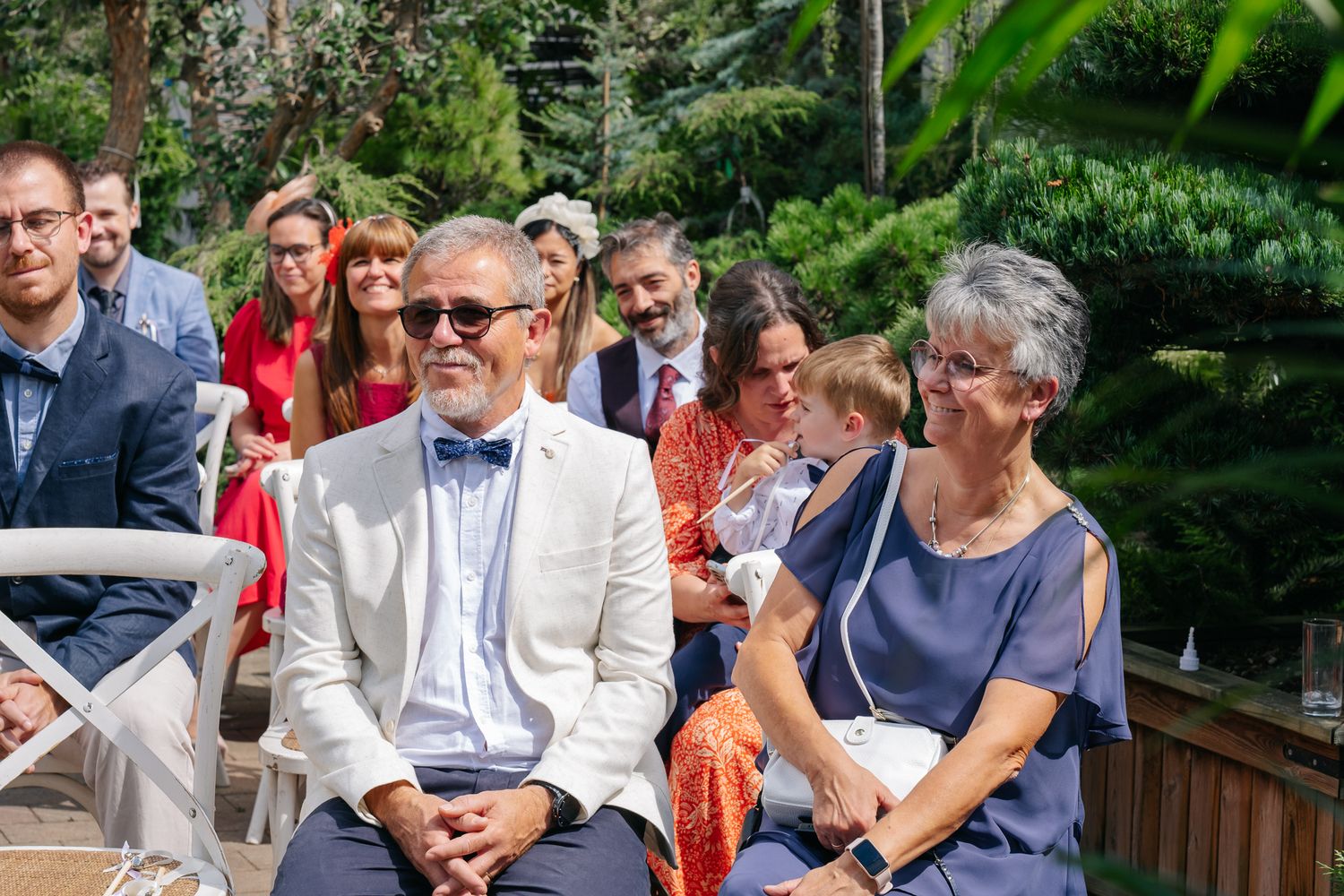 Smiling wedding guests seated outdoors, man in white blazer and bow tie beside woman in blue dress, lush greenery background.