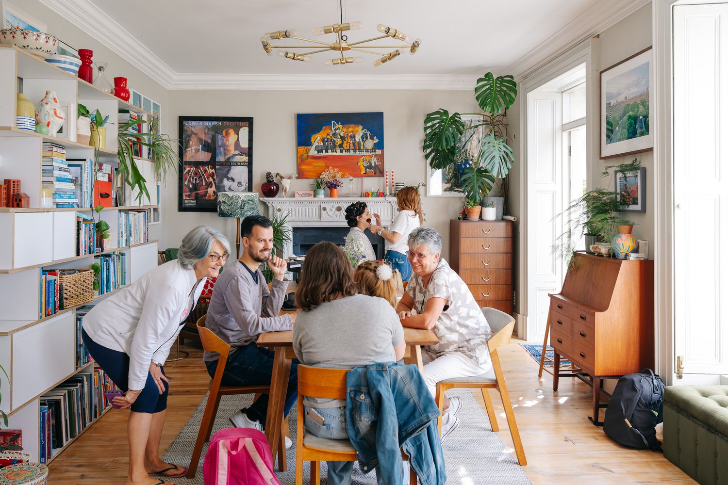 A diverse group of people gather around a dining table in a bright, art-filled living room with bookshelves and warm decor.