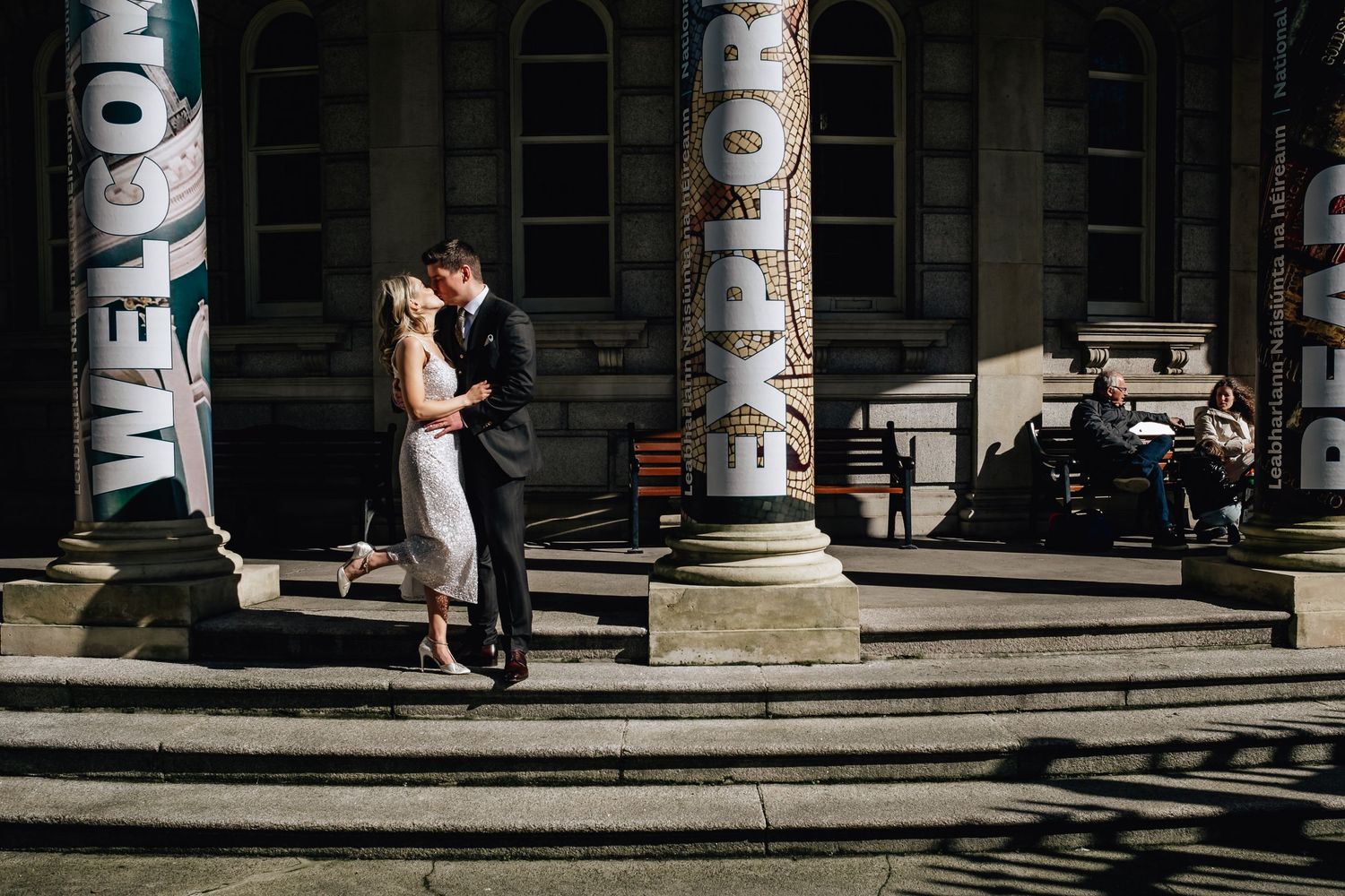 Couple embraces on stone steps between classical columns in dramatic lighting.