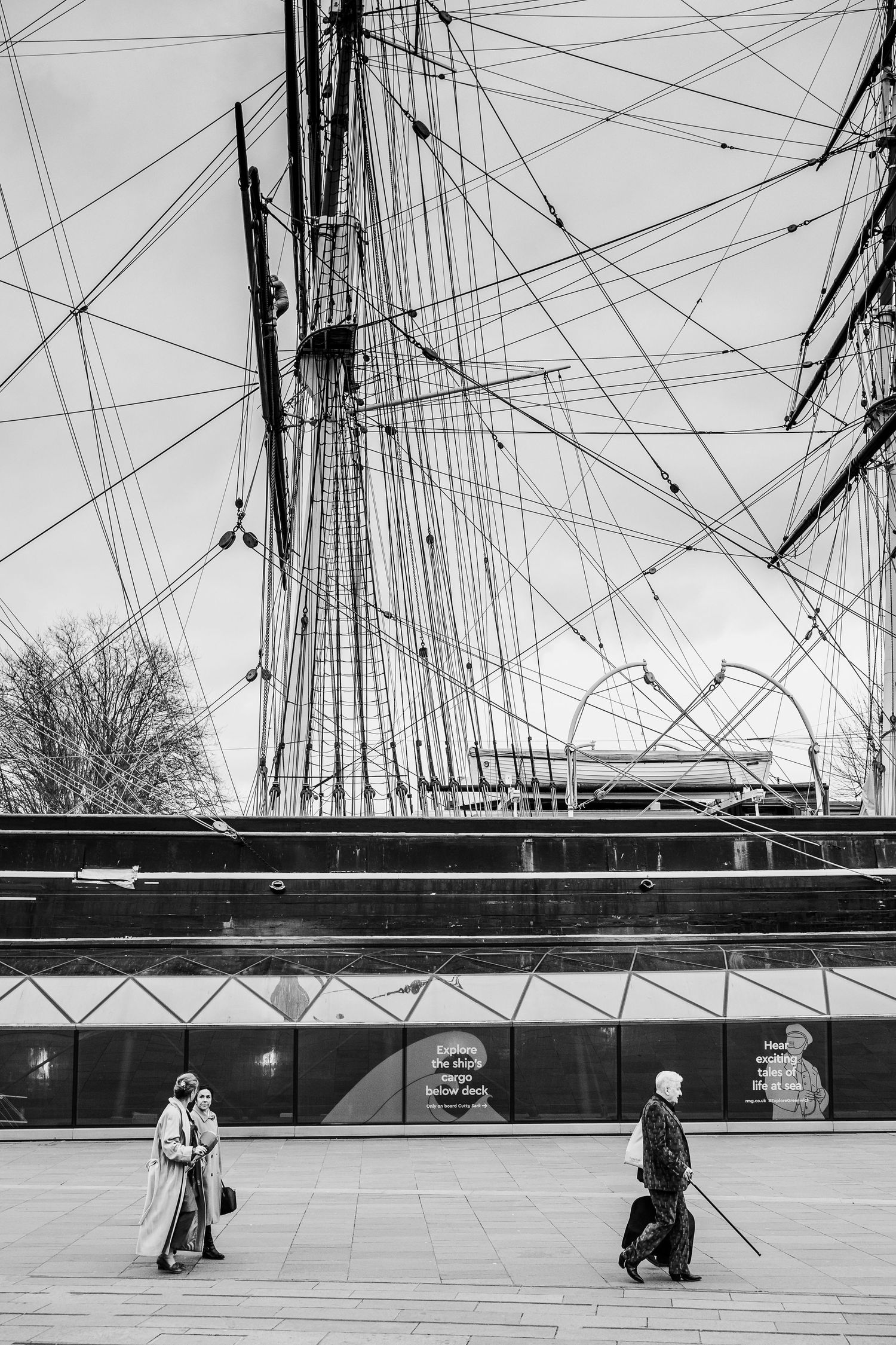 Black and white image of people walking beneath tall ship rigging and masts.