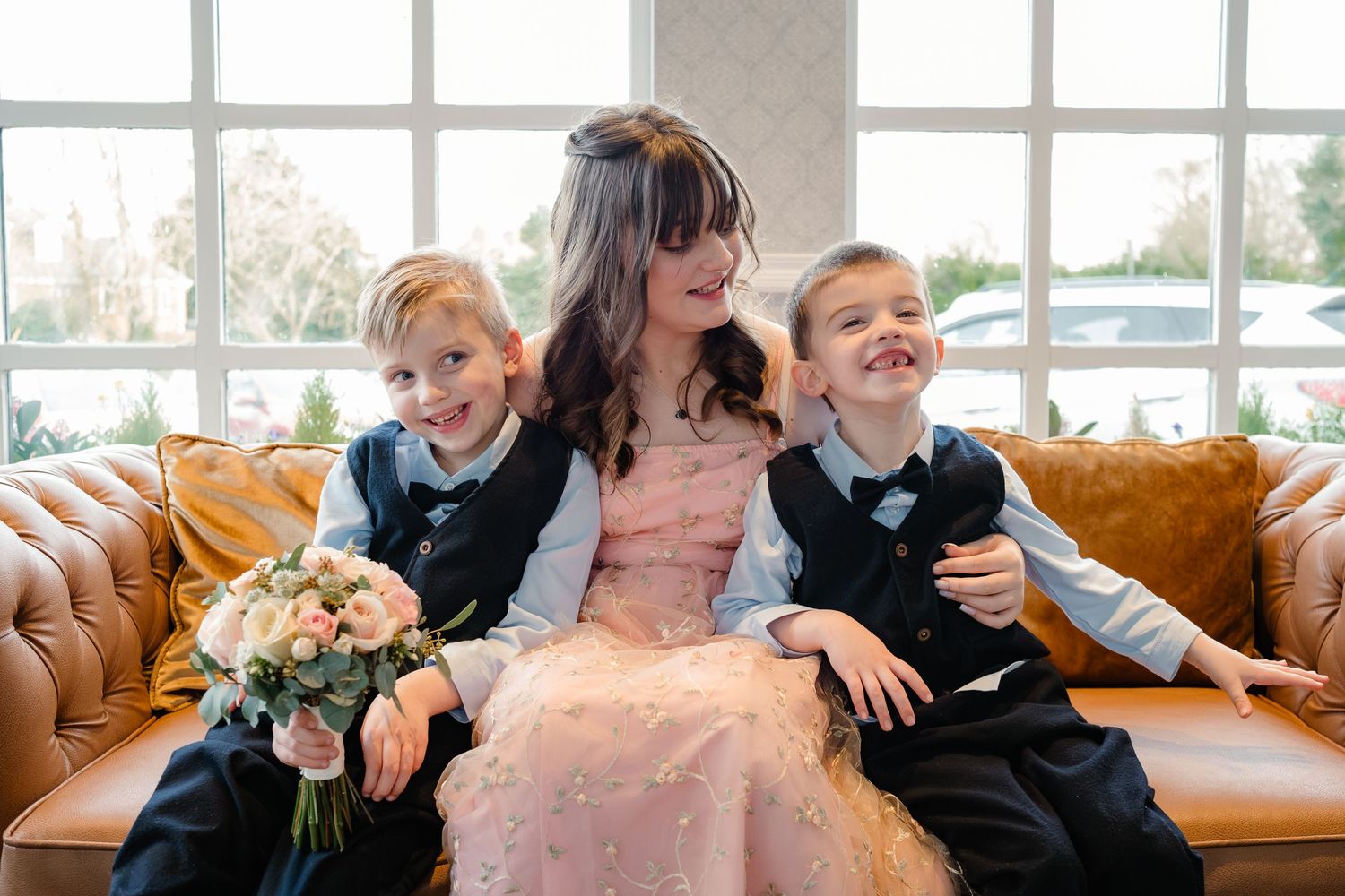 A joyful family moment on a tan leather couch features formal attire and floral bouquets during a special occasion.