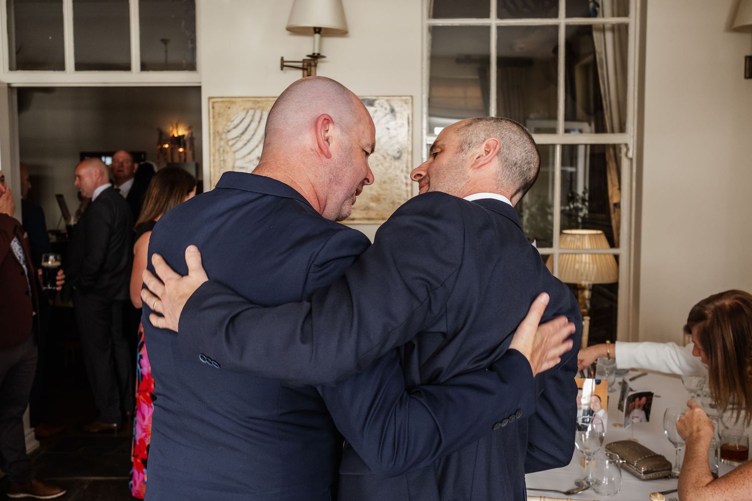 Two men in navy suits share a warm embrace at a formal wedding reception dinner.