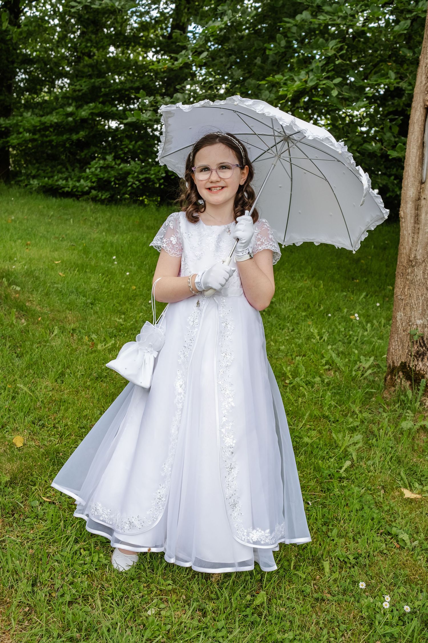 Girl in white First Communion dress, gloves, and parasol stands on green grass with trees in background.