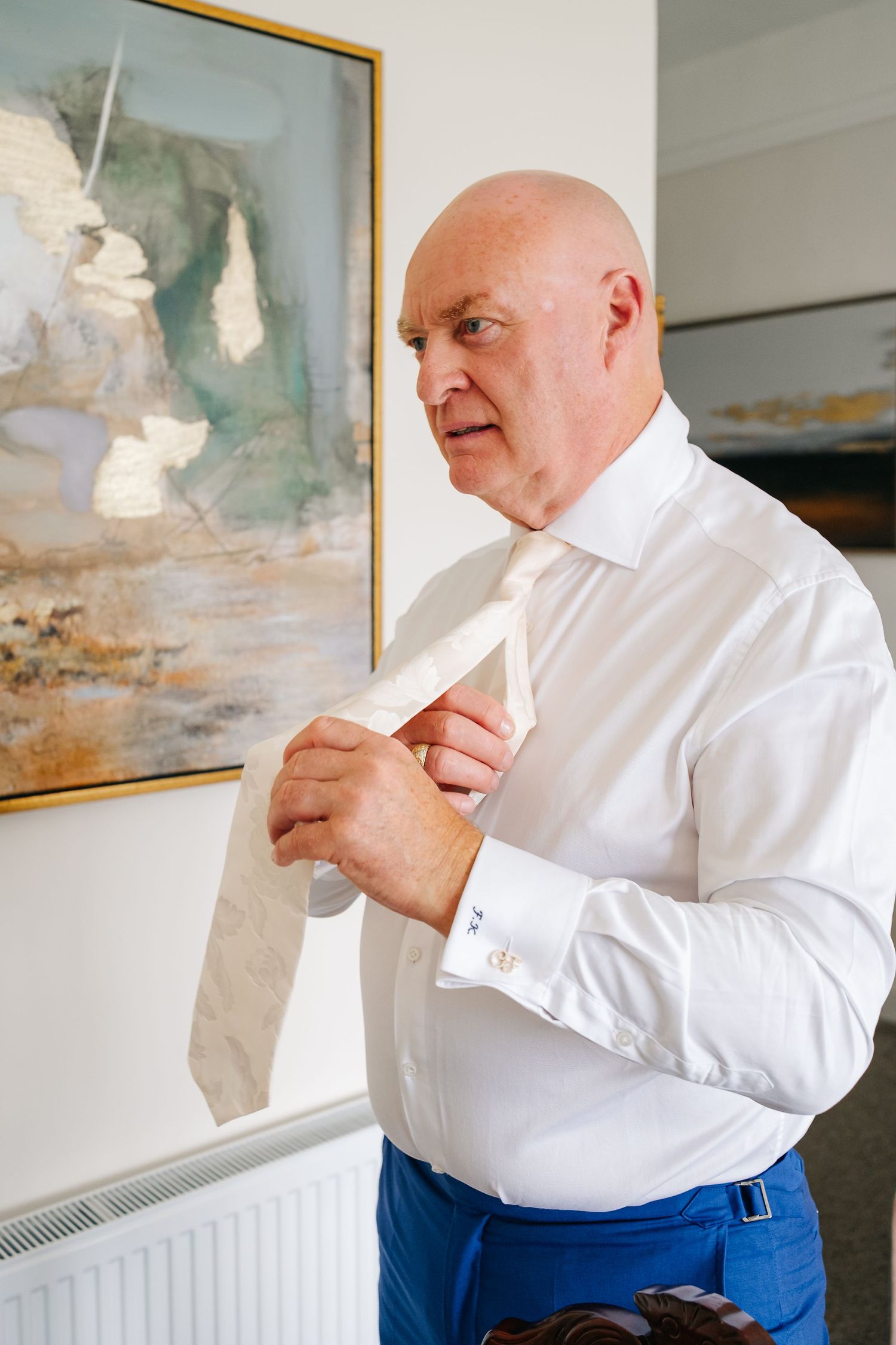 Bald man in white shirt adjusting a cream tie, standing indoors before a wedding ceremony.
