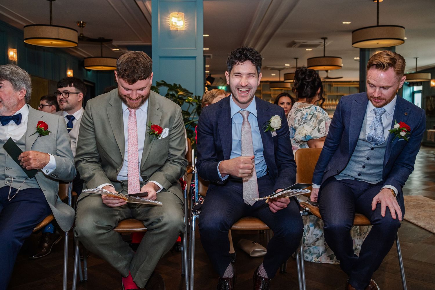 Groomsmen in suits reading wedding programs while seated at an indoor wedding ceremony venue.