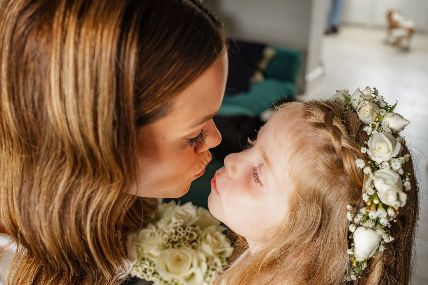 A bride and young flower girl share a tender nose-to-nose moment, girl wearing a floral crown holding white roses.