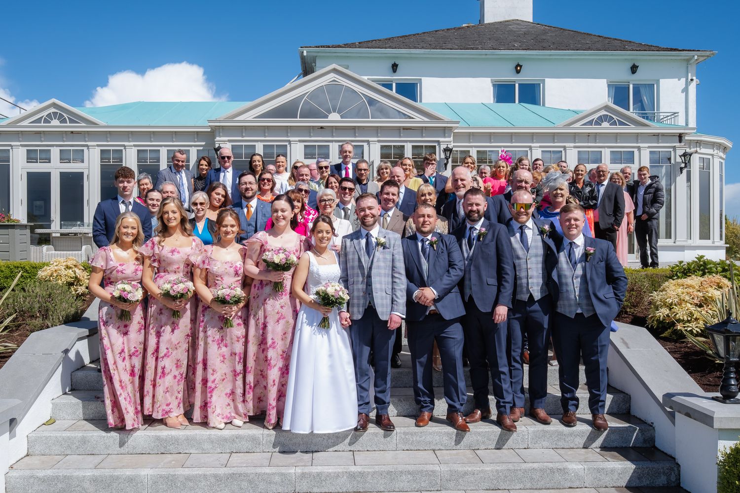 Large wedding party group photo outside elegant venue on sunny day, bride in white, bridesmaids in pink floral dresses.