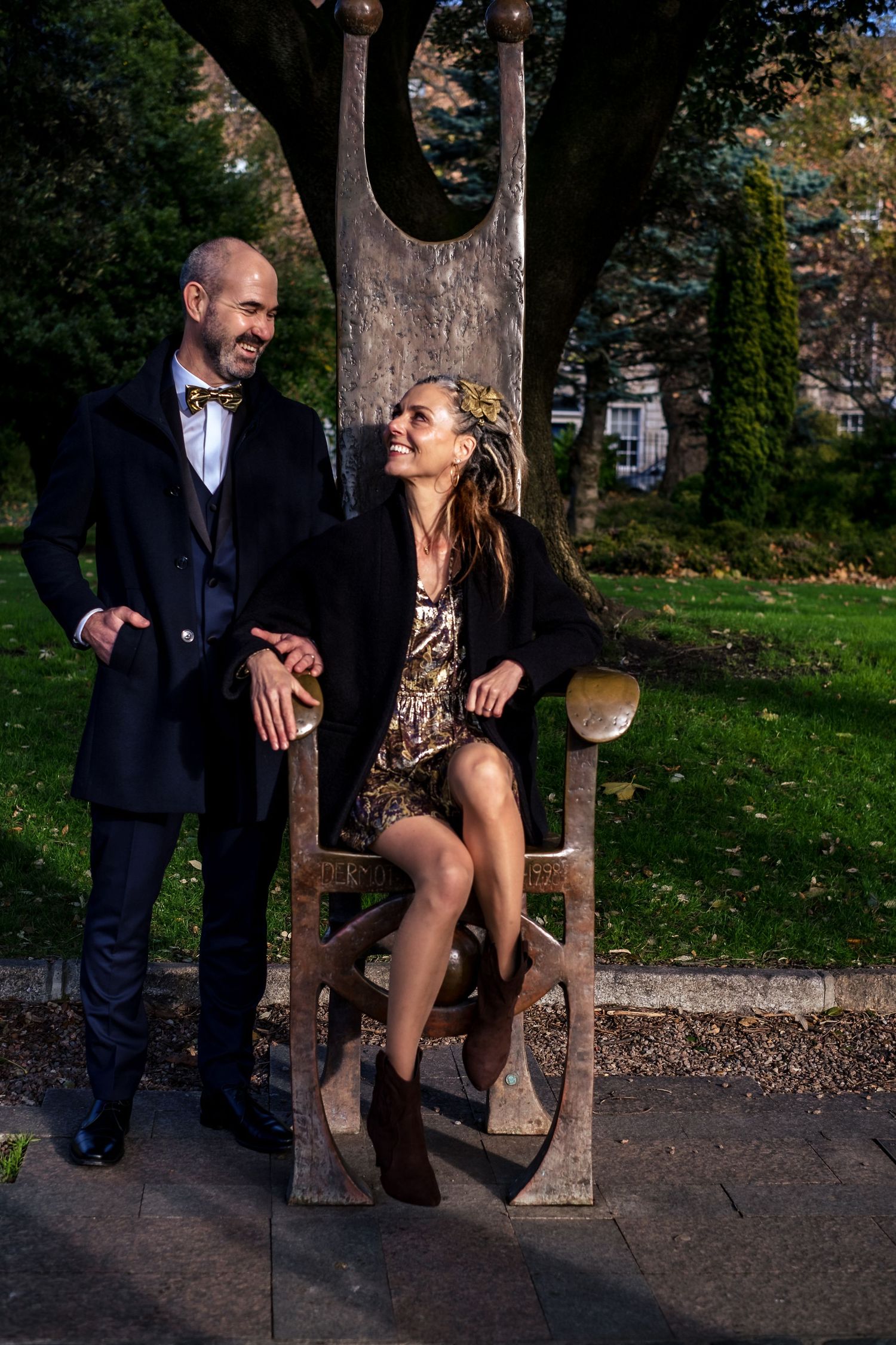 Couple smiling at each other in a park, man in suit with bow tie, woman in sequin dress seated on wooden chair.