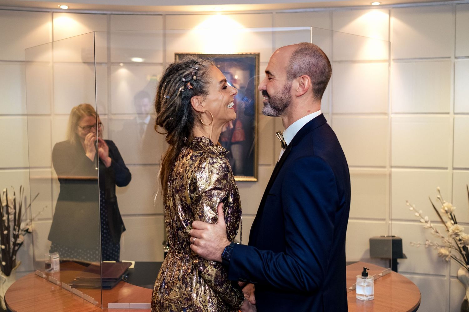 A woman in a gold dress and a man in a navy suit share an intimate moment at an elegant indoor event.