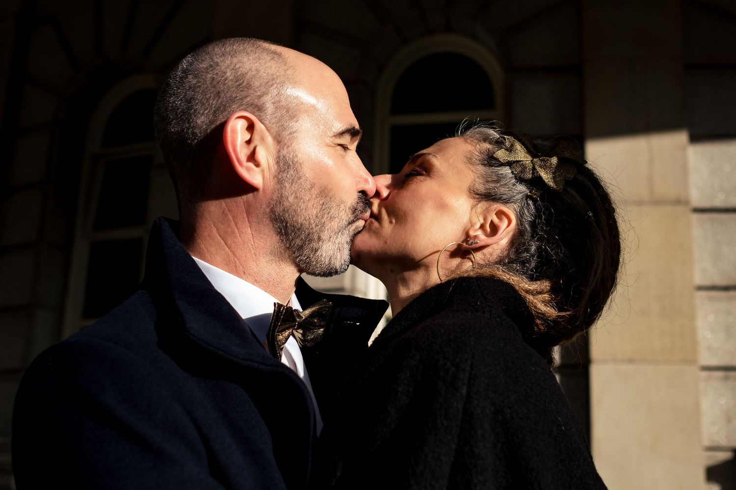 A couple shares a romantic kiss outdoors in warm sunlight, dressed in elegant attire.