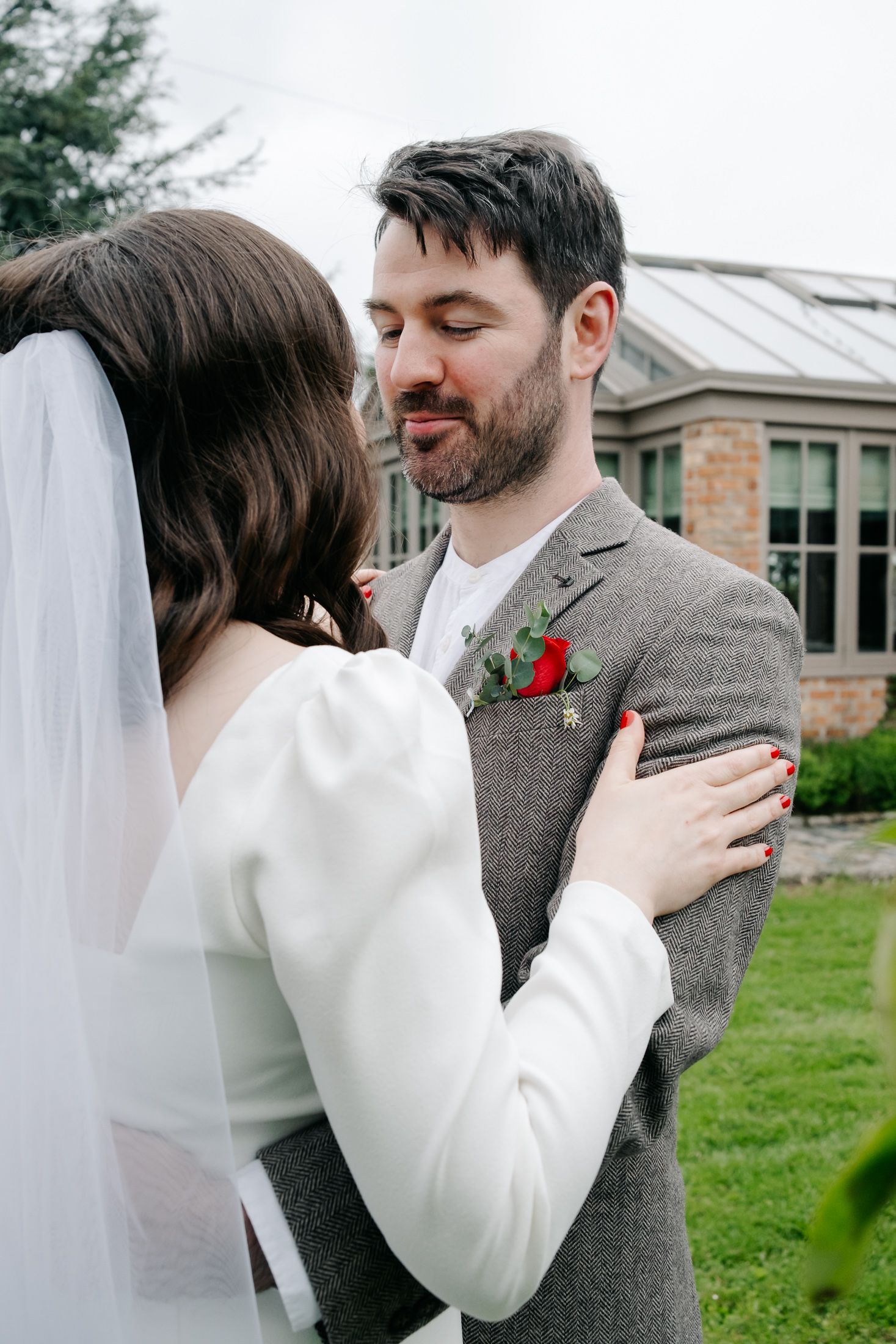 A groom in a gray suit with red boutonniere embraces his bride in a white dress and veil outdoors.