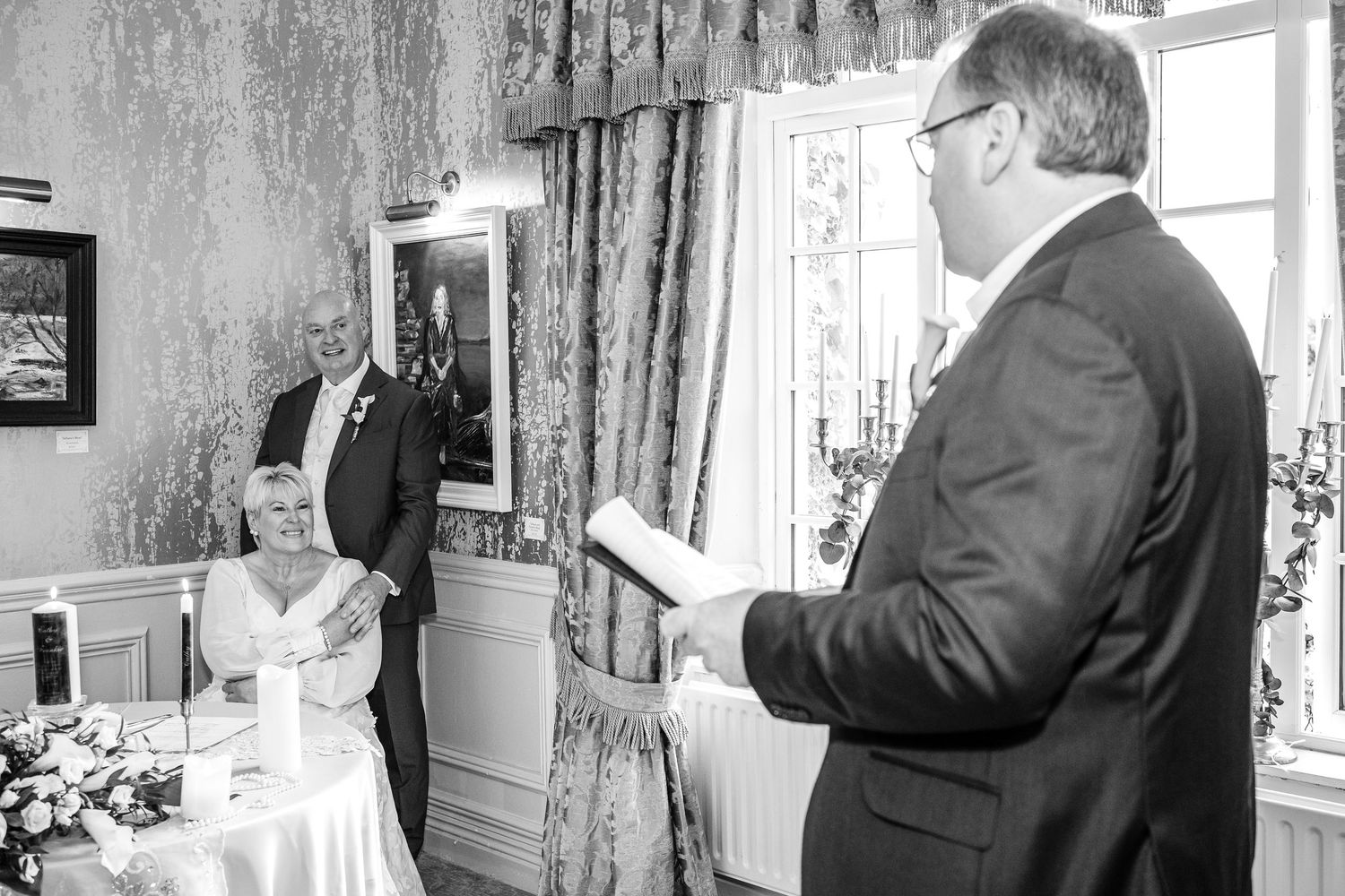 Black and white photo of a wedding officiant reading vows as the bride and groom listen attentively.