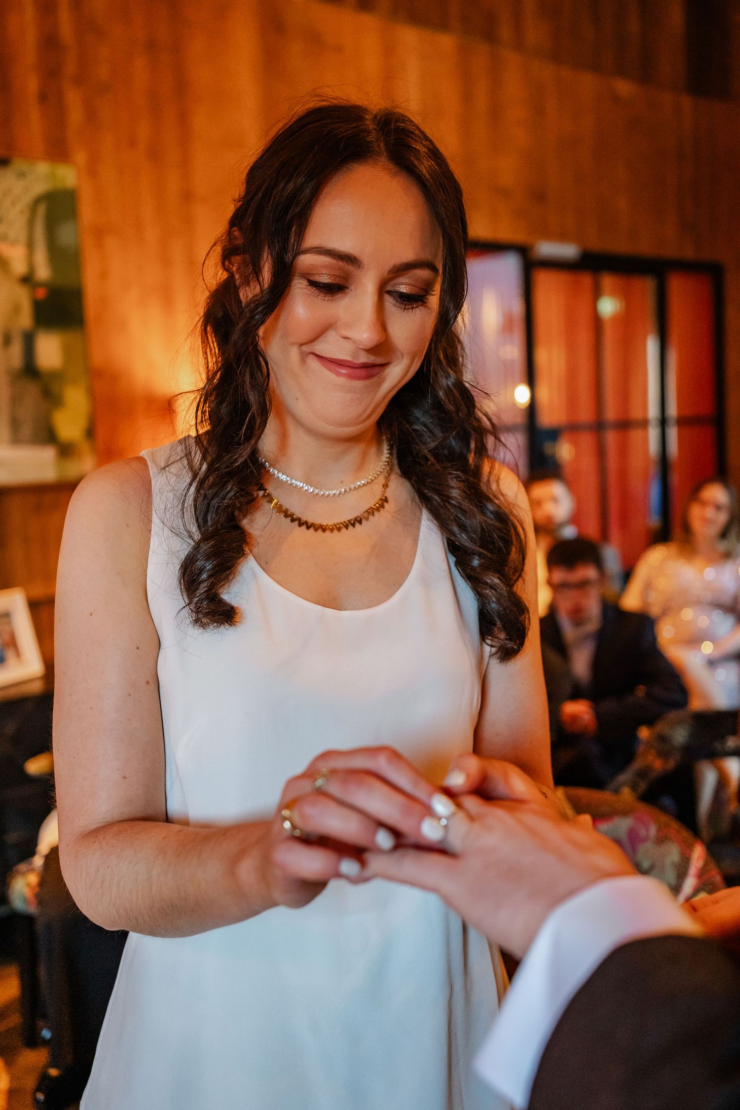 Bride smiling while placing wedding ring on groom's finger during indoor ceremony.