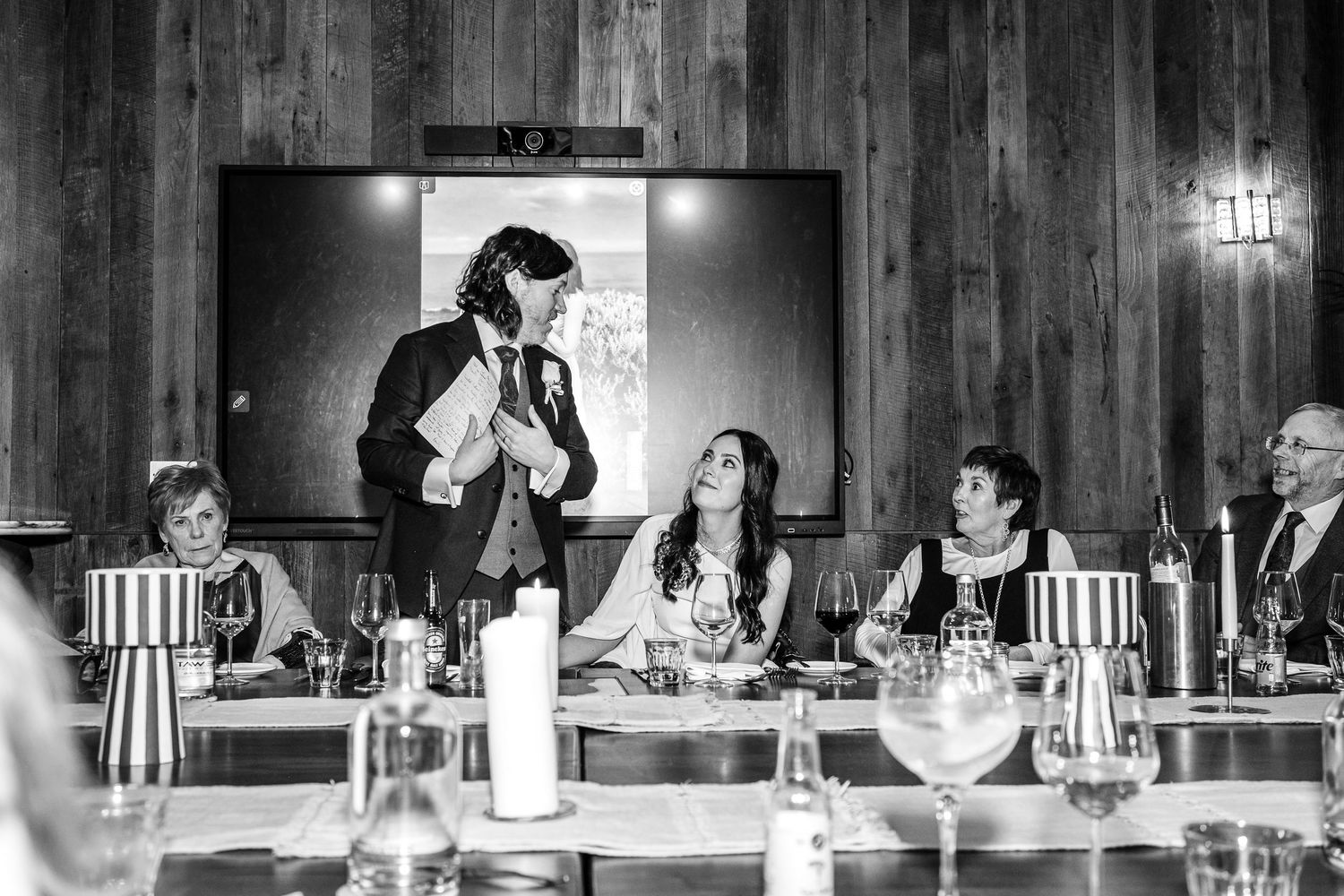 Black and white wedding reception photo of groom giving speech at head table with seated guests and candles.