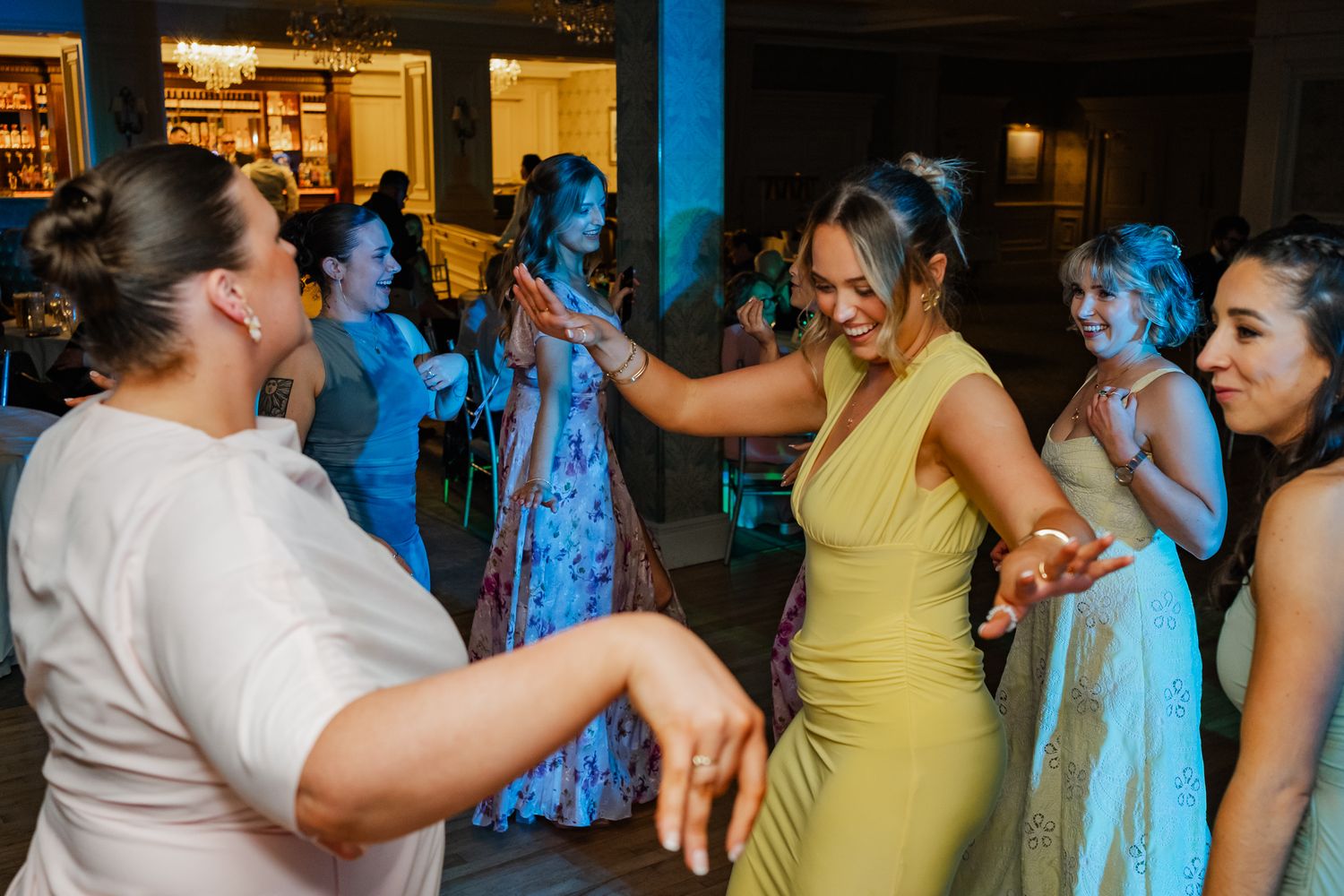 Women in elegant dresses dancing joyfully at a wedding reception party under blue lights.