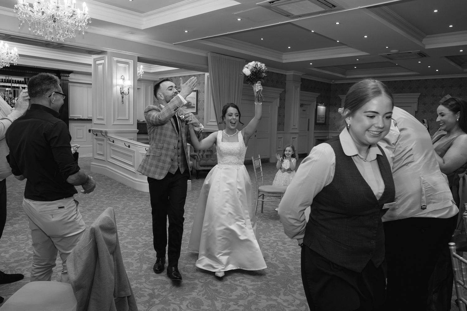 Black and white wedding photo of bride holding bouquet aloft while guests celebrate on dance floor.