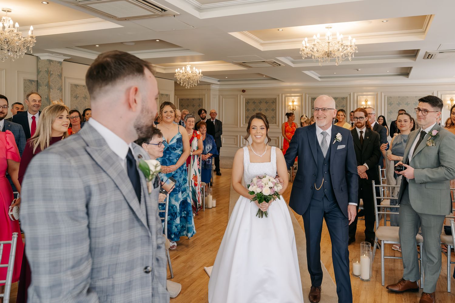 Bride walks down the aisle with her father toward the groom during an elegant wedding ceremony in a grand venue.