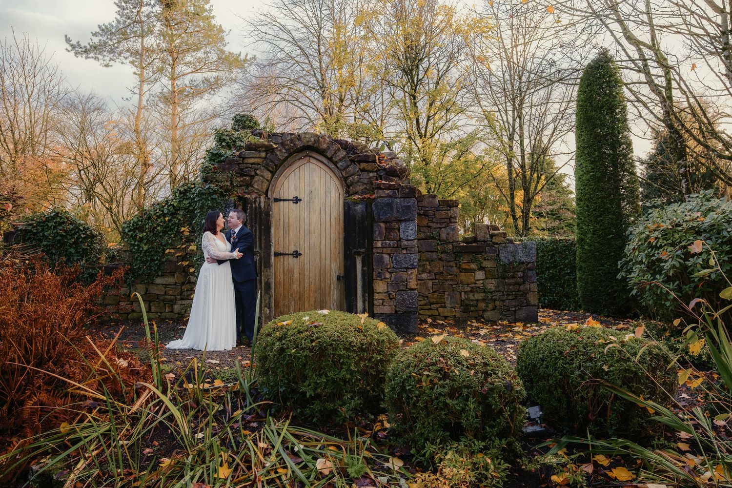 Bride and groom embracing in an autumn garden beside a rustic stone wall with a wooden arched door.