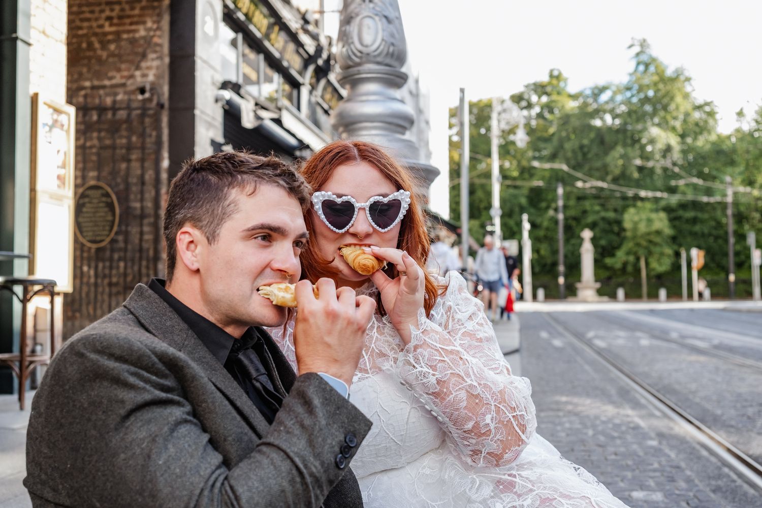 Bride in lace dress and heart sunglasses eating pastry with groom on urban street corner.