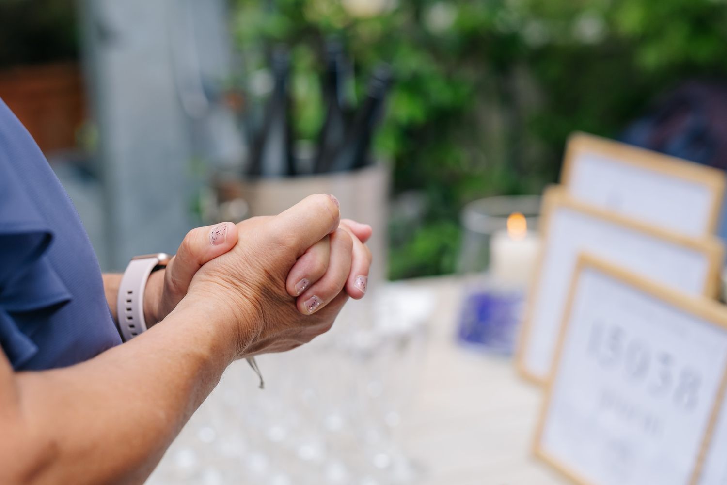 Close-up of hands clasped together at an outdoor wedding reception with candles and table numbers in background.