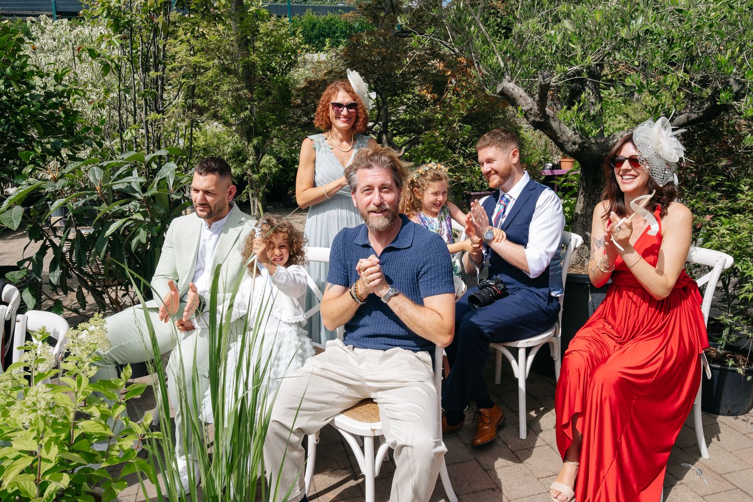 Group of guests celebrating outdoors at a sunny garden wedding ceremony, woman in red dress clapping joyfully.