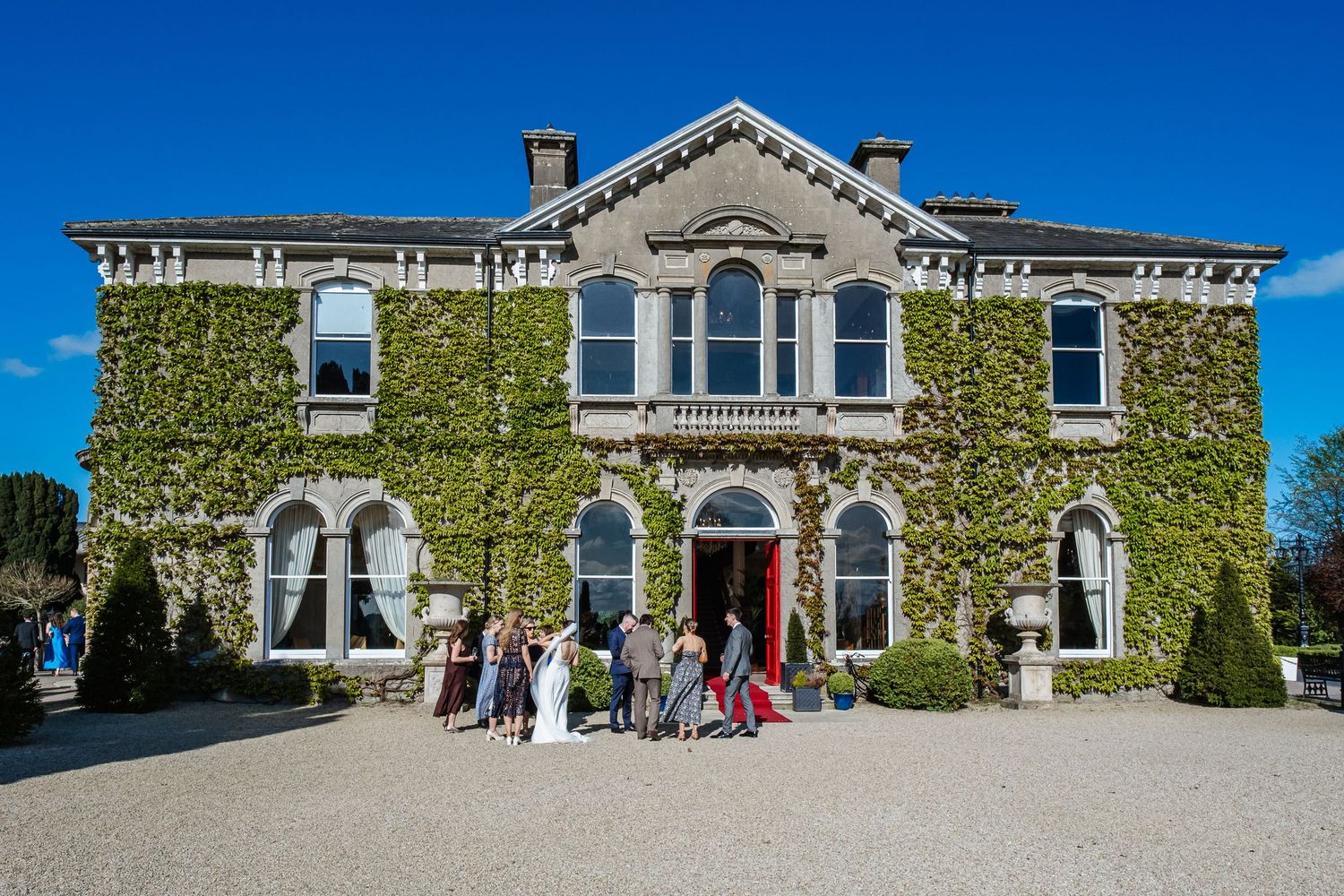 Panoramic view of historic mansion with ivy-covered walls and elegant architecture against bright blue sky on sunny day.